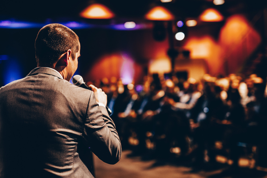 Palestrante conduzindo uma apresentação enquanto a plateia acompanha atentamente ao fundo